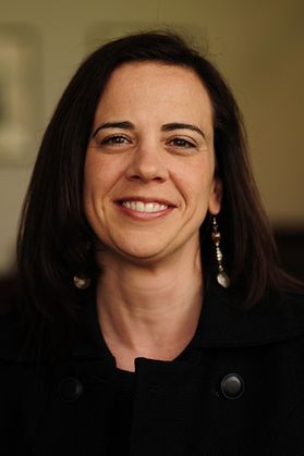 A woman with long dark hair wears a dark shirt and long earrings while smiling for the camera
