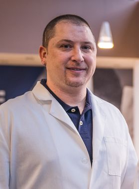 Headshot of WVU School of Dentistry professor Christopher Waters. He is pictured inside wearing his white lab coat over a blue golf shirt. He has very short dark hair and a light goat tee. 