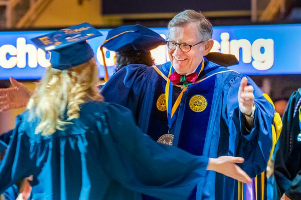 man with grey hair and glasses in a graduation gown greets female student in cap and gown on stage