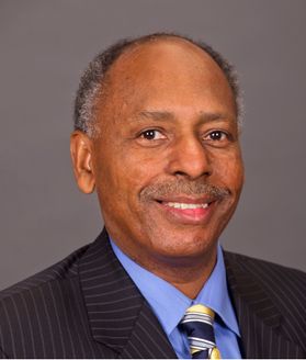 Headshot of retired WVU dean Dana Brooks. He is pictured in front of a gray background wearing a dark colored pinstriped suit, blue dress shirt, and blue and yellow patterned tie. He has short black and gray hair and a faint gray mustache. 