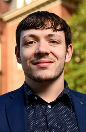 Headshot of WVU scholarship recipient Matthew Kinzer. He is pictured outside with Woodburn Hall behind him. He is wearing a navy blue jacket over a dark colored shirt. He has short, brown hair and a light beard. 