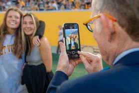 An older man with glasses takes a photo on a phone of two young girls on a football field