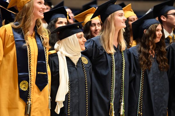 Four graduates are lined up with their armed links. One is wearing a gold gown. The others are in navy blue gowns. They are looking up and to the left and smiling.